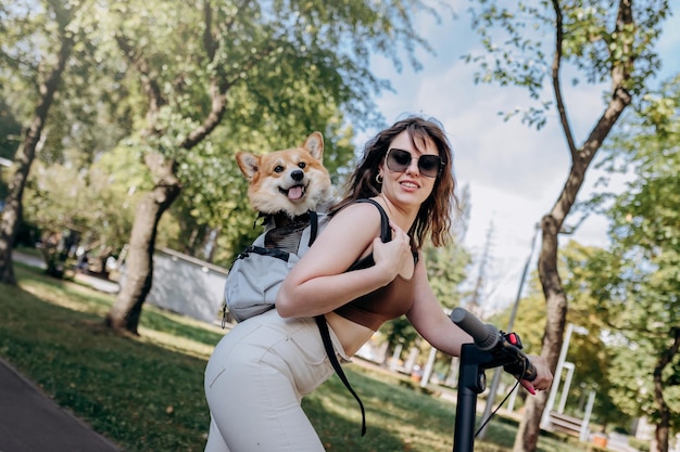 Gelukkig lachende vrouw reiziger rijdt op haar elektrische scooter in stadspark met hond Welsh Corgi Pembroke in een speciale rugzak