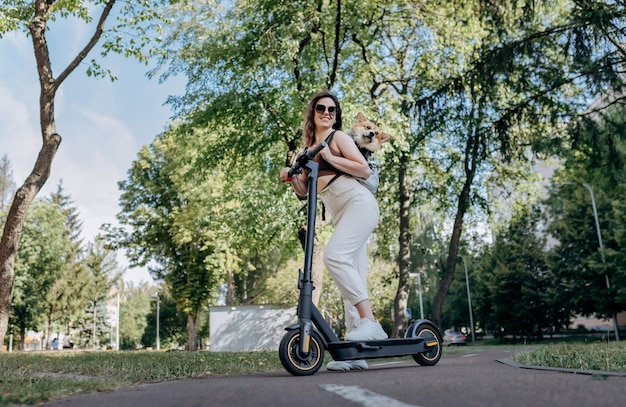 Gelukkig lachende vrouw reiziger rijdt op haar elektrische scooter in stadspark met hond Welsh Corgi Pembroke in een speciale rugzak