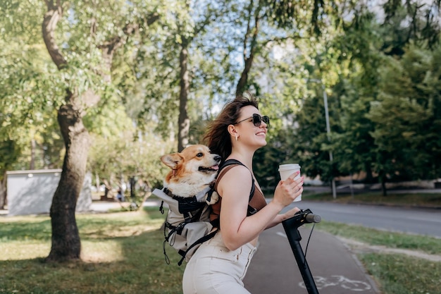 Gelukkig lachende vrouw reiziger staat met electro scooter en koffie drinken in stadspark met hond Welsh Corgi Pembroke in een speciale rugzak