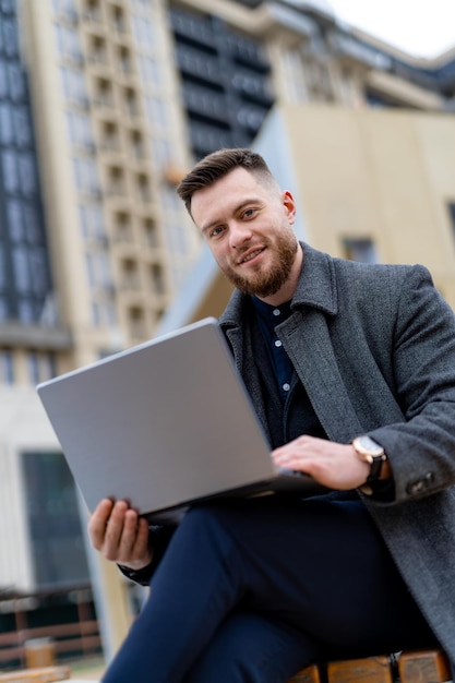 Gelukkig lachende zakenman in jas werken en kijken met laptop op moderne brede straat.