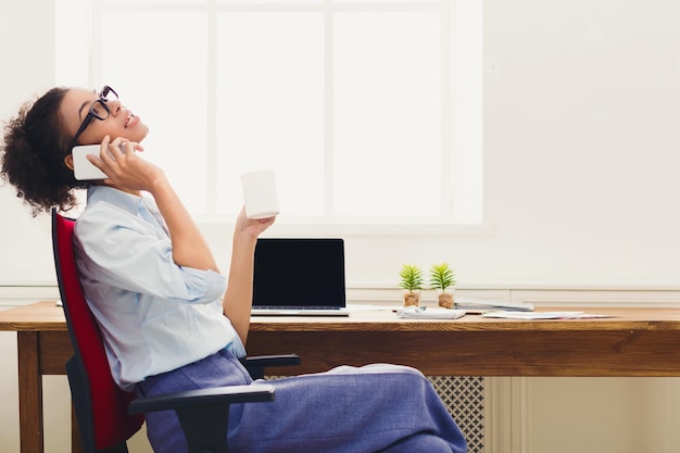 Gelukkig lachende zakenvrouw aan het werk praten aan de telefoon, zittend op haar werkplek op kantoor en koffiepauze, kopieer ruimte, zijaanzicht