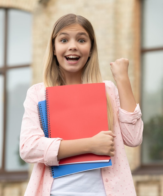 Gelukkig leerling meisje met boeken die een winnend gebaar maken buitenshuis backtoschool