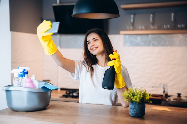 Gelukkig leuke jongedame in gele handschoenen neemt selfie tijdens het schoonmaken van haar keuken thuis met wasmiddelen