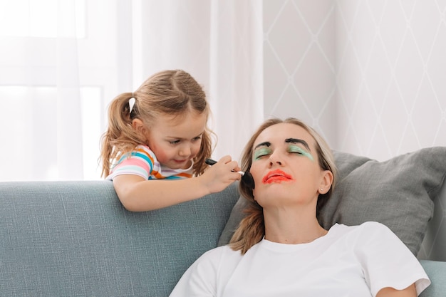 Gelukkig liefdevol gezin. Jonge moeder slapen op de bank thuis terwijl kind meisje gezicht gekleurde aquarellen verf. Grappige make-up. plezier hebben. Prank