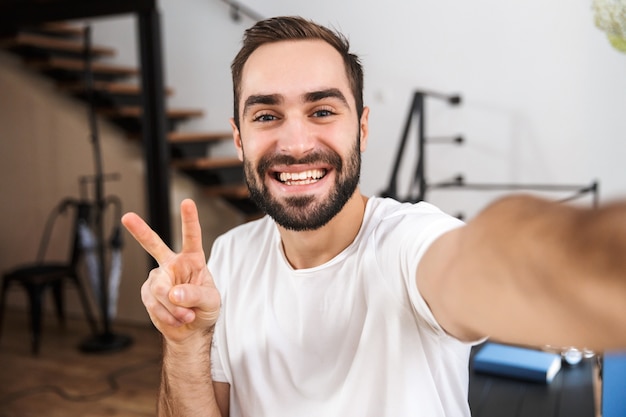 Gelukkig man een selfie te nemen zittend in de keuken thuis, vredesgebaar