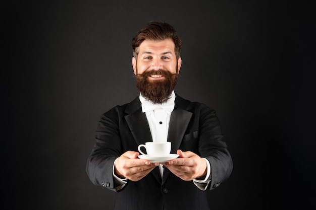 Gelukkig man in smoking vlinderdas met koffiekopje. ober in formalwear die koffie serveert op zwarte achtergrond