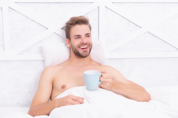 Gelukkig man koffie drinken uit beker in witte slaapkamer op bed