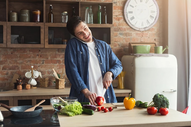 Gelukkig man koken gezond voedsel en praten op smartphone in loft keuken thuis op zonnige dag. Groentesalade bereiden, ruimte kopiëren