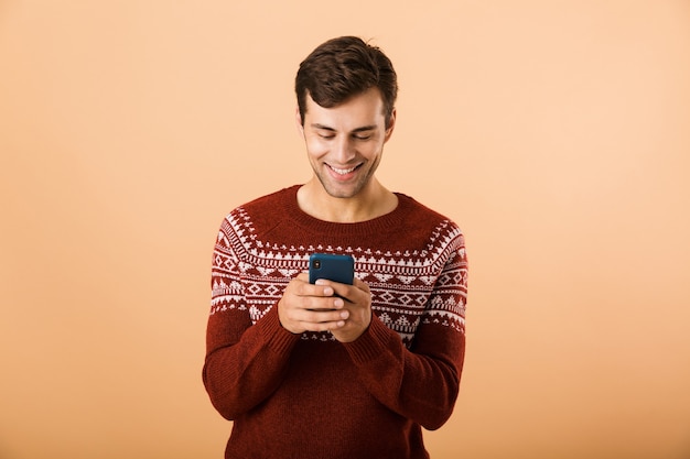 Gelukkig man met stoppels dragen gebreide trui met behulp van smartphone, geïsoleerd over beige muur