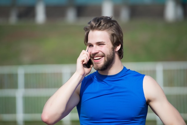 Gelukkig man praten op mobiele telefoon op zonnige buiten, communicatie. Bebaarde man glimlach met smartphone, moderne leven. Sportman in blauwe sport tshirt, mode. Sportmode en een gezonde levensstijl.