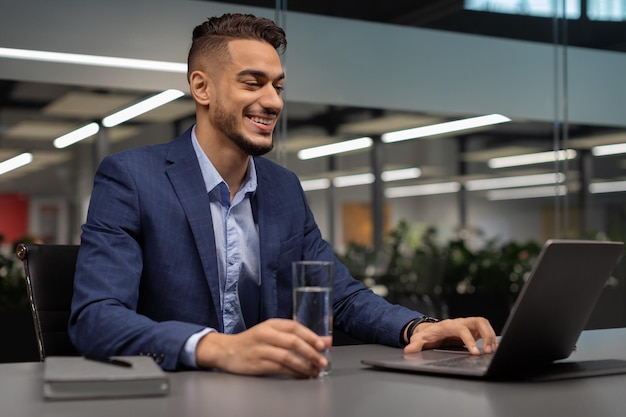 Gelukkig manager drinkwater tijdens het werken op laptop