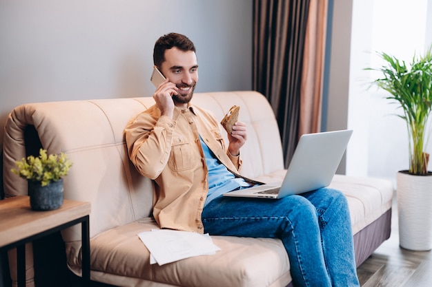 Gelukkig mannelijke freelancer zit op de bank in zijn woonkamer en spreekt met een klant met behulp van mobiele telefoon kijken naar taak op laptop scherm