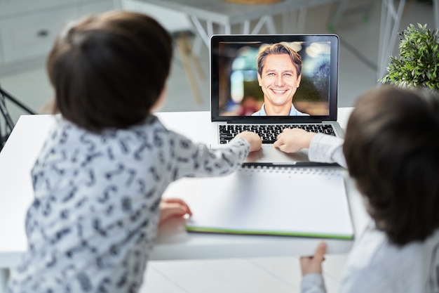 Gelukkig mannelijke leraar lacht naar zijn kleine studenten tijdens online les voor kinderen. Twee Spaanse jongens die laptop gebruiken terwijl ze aan tafel zitten. Afstandsonderwijs voor kinderen. Focus op laptopscherm