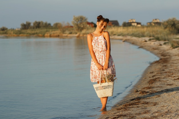 gelukkig meisje bij zonsondergang aan zee met een strozak en een hoed gaat op blote voeten. De achtergrond is wazig.