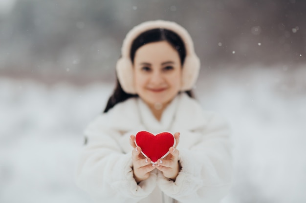 Gelukkig meisje dat in warme witte kleren rood hart op besneeuwde achtergrond houdt. Valentijnsdag.