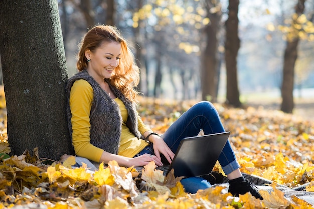 Gelukkig meisje dat op laptop werkt die onder een boom zit in het herfstpark met gele bladeren