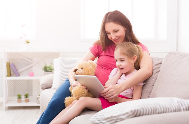 Gelukkig meisje en haar moeder met behulp van digitale tablet, zittend op de bank thuis. Moeder laat haar dochter foto's zien, tekenfilm, ontwikkelingsgames spelen, ruimte kopiëren