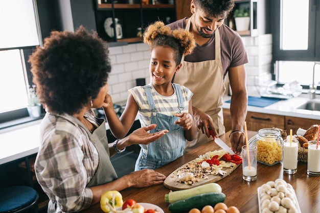 Foto gelukkig meisje en haar mooie ouders glimlachen terwijl ze thuis in de keuken koken