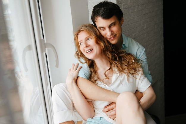 Gelukkig meisje en man knuffelen in de buurt van venster in huis. Witte en blauwe kleding. Valentijnsdag.