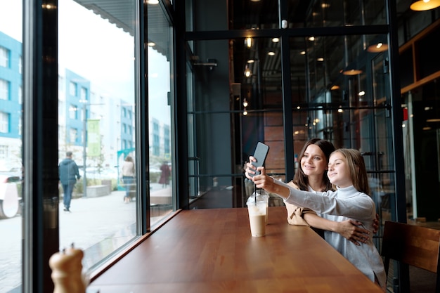 Gelukkig meisje haar armen strekken met smartphone tijdens het maken van selfie met moeder in café door raam