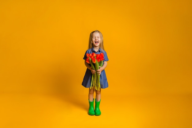 Gelukkig meisje in een blauwe denimjurk en groene rubberen laarzen met rode tulpen op een gele muur