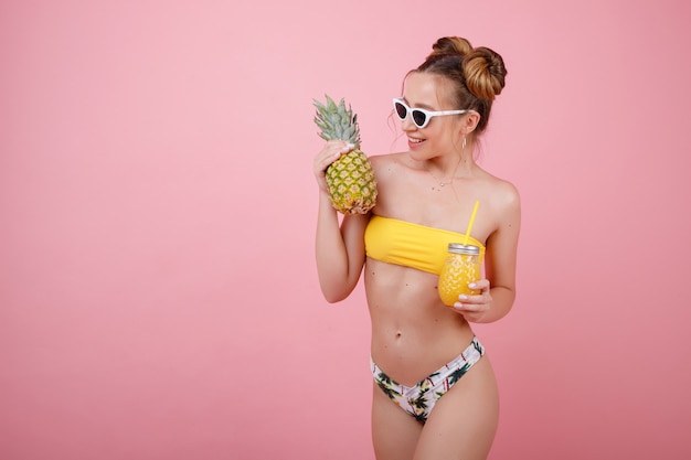Gelukkig meisje in een gele zwembroek en glazen met sap in een glas en ananas in haar handen op een lichte zomer ruimte