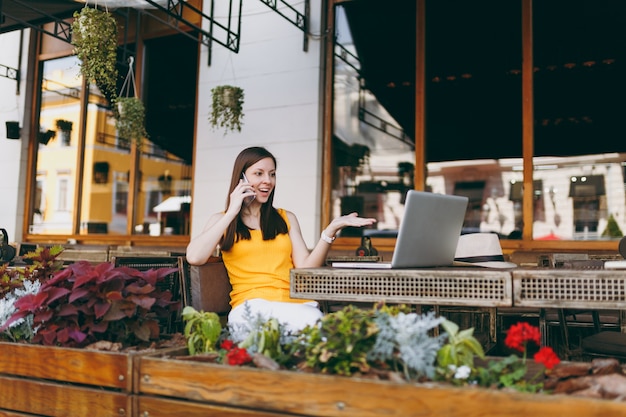 Gelukkig meisje in openlucht straatcafé zittend aan tafel met laptop pc-computer, praten op mobiele telefoon, aangenaam gesprek voeren, in restaurant tijdens vrije tijd