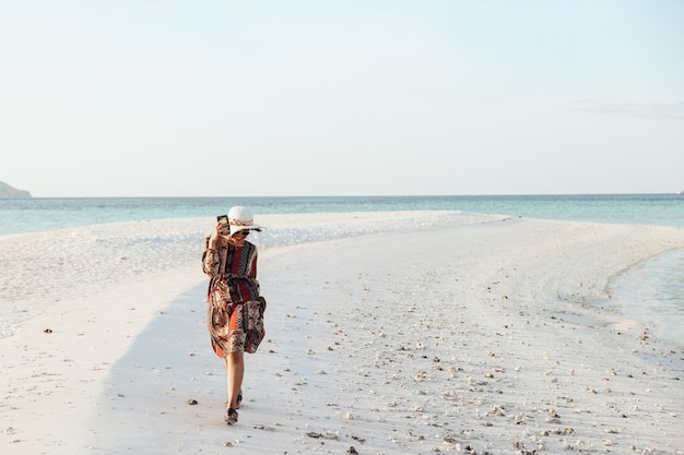 Gelukkig meisje in zomerhoed en zonnebril die een smartphone vasthoudt terwijl ze rennen en spelen op het strand