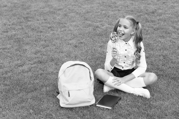 Gelukkig meisje kind in uniform met schooltas en boek geniet van het eten van smakelijke lolly zittend op gras