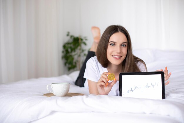 Gelukkig meisje met cryptomunt en tablet met financiële afbeelding, liggend in bed