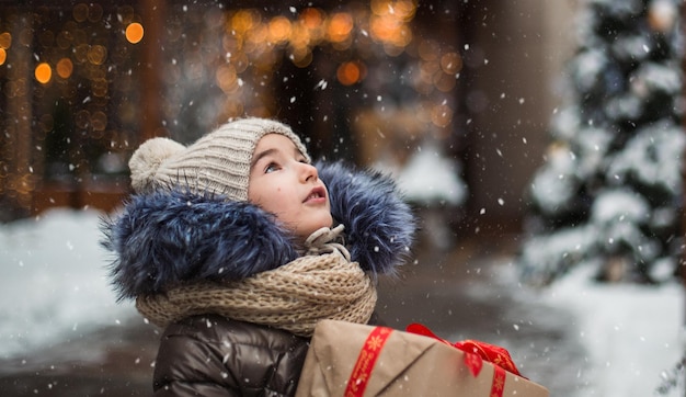 Gelukkig meisje met een geschenkdoos voor Kerstmis vangt sneeuwvlokken met zijn tong op een stad in de winter met sneeuw op de feestelijke markt met versieringen en verlichting. Warme kleding, gebreide muts, sjaal en bont.