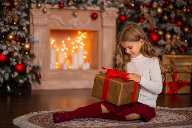 Gelukkig meisje met een nieuwjaarsgeschenk in handen op een achtergrond van een open haard en een kerstboom in de lichten