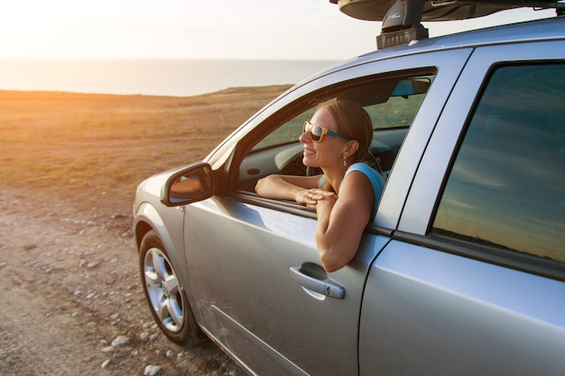 Gelukkig meisje met zonnebril kijkt uit het raam van haar auto op een prachtige zonsondergangachtergrond