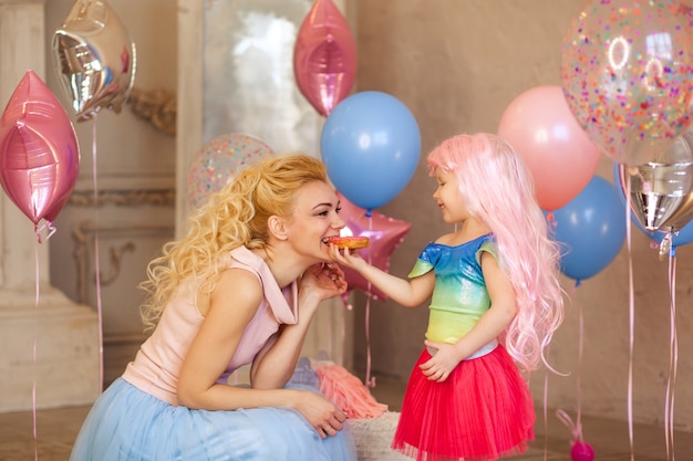 Gelukkig meisje van 3-4 jaar in een roze pruik geeft een lekkere donut aan haar moeder, baby's verjaardag