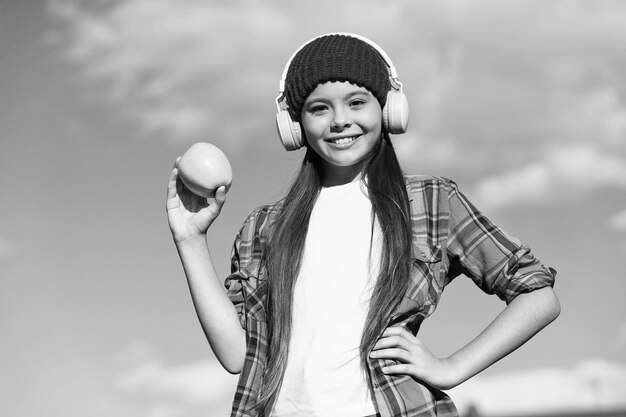 Gelukkig meisjeskind in casual modestijl, luister naar muziek in moderne koptelefoons met verse, smakelijke appel, zonnige hemelvitamine