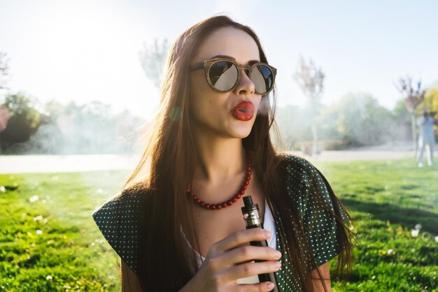 Gelukkig mode lachende vrouw in zonnebril roken vape op straat, rook