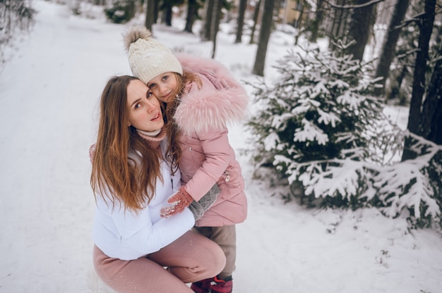 Gelukkig moeder en klein schattig meisje in roze warme uitloper wandelen met plezier en knuffelen in besneeuwde witte koude winter naaldbos met sparren bossen buitenshuis