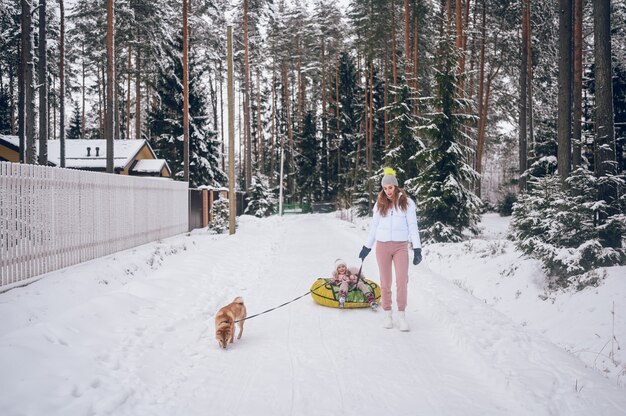 Gelukkig moeder en schattig meisje in roze warme uitloper wandelen plezier rijdt opblaasbare sneeuw buis met rode shiba inu hond in besneeuwde witte koude winter forest buitenshuis