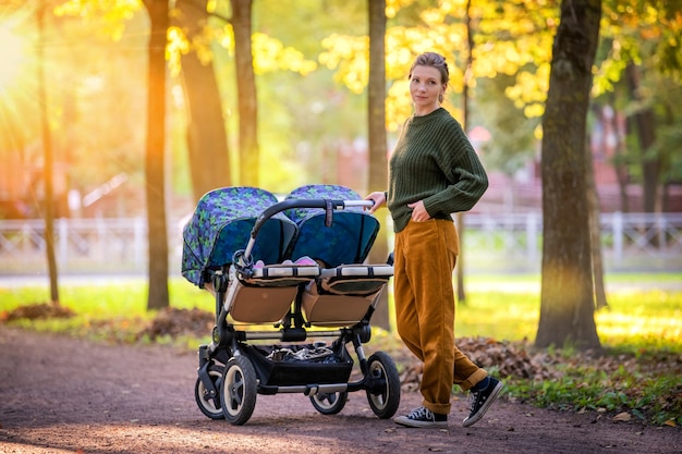 Gelukkig moeder wandelen in herfst park met tweeling kinderwagen