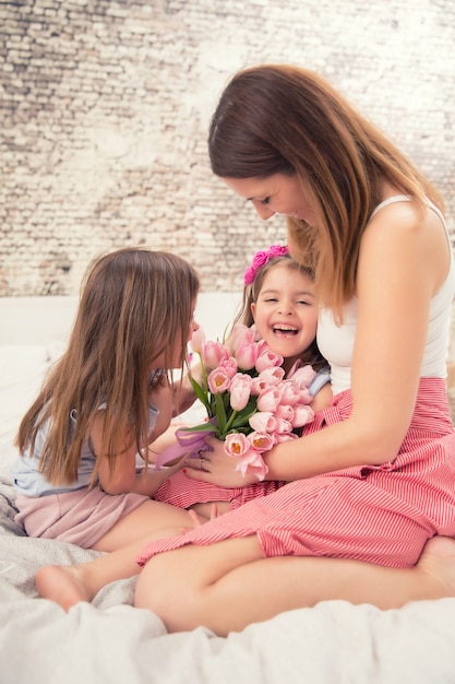 Gelukkig moederdagconcept. Moeder met twee schattige jonge dochters tweeling op het bed in de slaapkamer en een boeket bloemen tulpen.