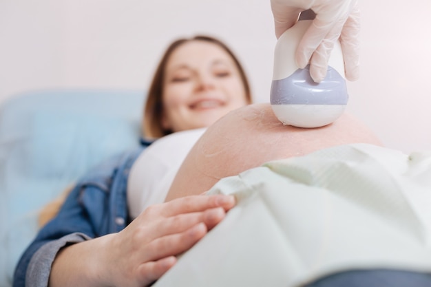 Gelukkig moment van moederschap. Jonge heerlijke geïnspireerde vrouw genieten van afspraak in de kliniek en het krijgen van ultrasone controle van haar buik terwijl arts met behulp van lineaire ultrasone sonde