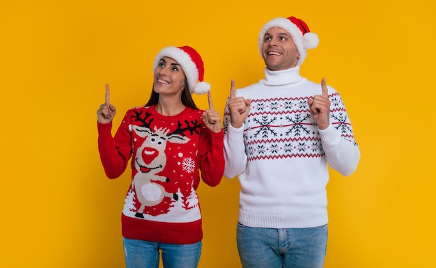 Gelukkig mooi glimlachend jong koppel verliefd in wintertruien en kerstmutsen terwijl ze naar boven wijzen en poseren geïsoleerd op gele achtergrond