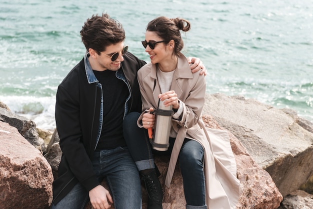 Gelukkig mooi jong stel met jassen op het strand wandelen, handen vasthouden, koffie drinken uit thermos