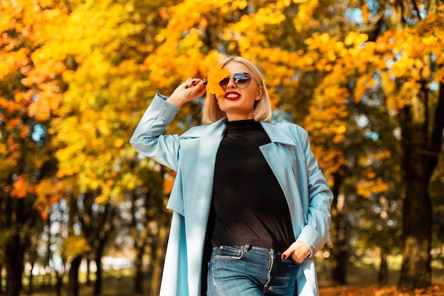 Gelukkig mooi lachend meisje in modekleding bedekt haar gezicht met geel herfstblad en loopt in het park tegen de achtergrond van felgekleurd herfstgebladerte