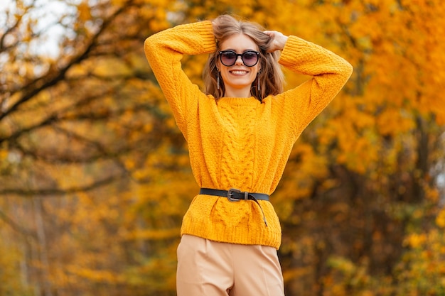 Gelukkig mooi tevreden jong meisje met een glimlach in modieuze zonbeschermingsbril in een gebreide gele trui loopt op het platteland met oranje herfstbladeren