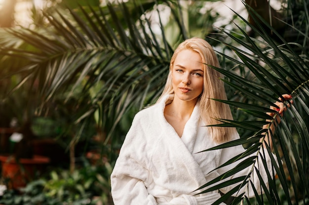 Gelukkig mooi vrolijk vrouwelijk model met een witte badjas die achter een palmblad staat, naar de zijkant kijkt en glimlacht. spa, behandeling, natuurlijke cosmetica, huidverzorging, cosmetologie concept.