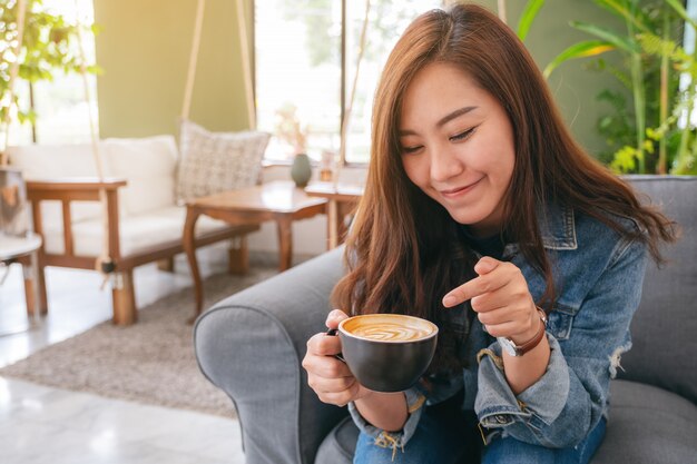 gelukkig mooie Aziatische vrouw bedrijf en wijzende vinger op een kopje warme koffie in café