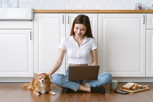 Gelukkig mooie Aziatische vrouw freelancer met behulp van laptopcomputer zittend op de vloer moderne keuken met haar mooie chihuahua hond zit naast thuis Werken vanuit huis Nieuw normaal en vriendelijk hondenconcept