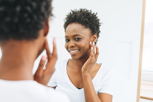 Gelukkig mooie jonge Afro-Amerikaanse vrouw in witte t-shirt zet oorbel tegen de spiegel