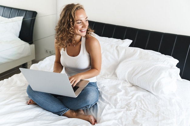 Gelukkig mooie jonge blonde harige vrouw zittend op bed, met behulp van laptopcomputer